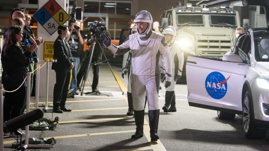 astronaut in white spacesuit waves to media behind a cordon, backdropped by nasa vehicles