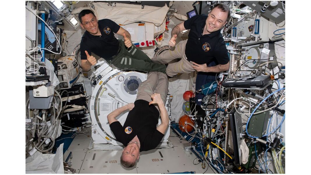 three men in black polo shirts float aboard the international space station.