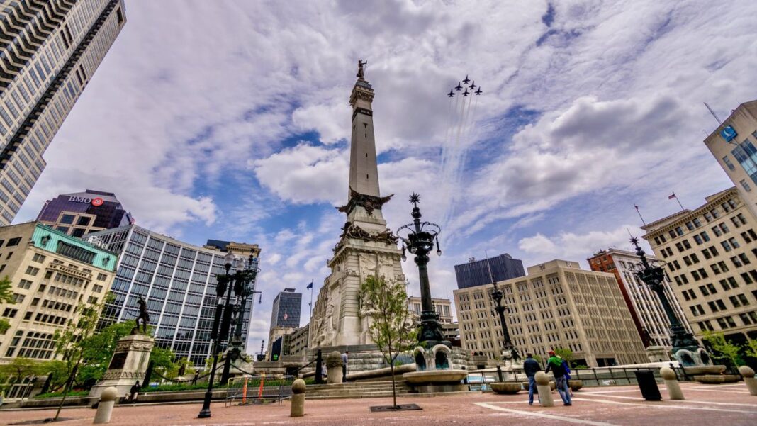 Six jets in formation fly through a cloudy blue sky over a city